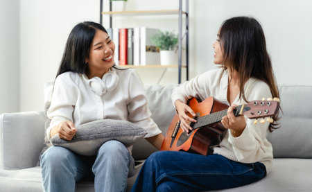 Two women are sitting on a couch, one of them playing a guitar. They are smiling and seem to be...