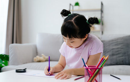 A young girl is sitting on a couch and drawing with color pencil. She is wearing a pink shirt and has her hair in pigtails. The scene is peaceful and calm, with the girl focused on her drawingの写真素材