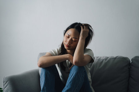 A lonely woman is sitting on a couch with her head in her hands. She is sad or upset.の写真素材