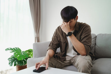 The young man placed his phone on the table and was sitting on the sofa in the living room. He looked tired after playing with his phone for a long time.の写真素材