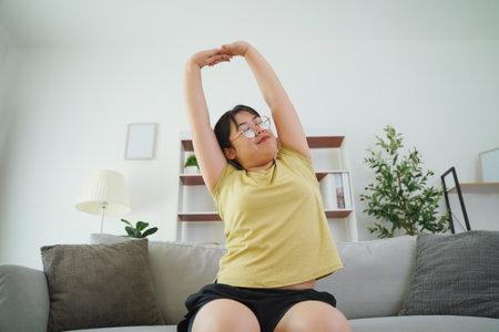 An Asian woman is stretching her muscles while sitting on a sofa in her living room at home.