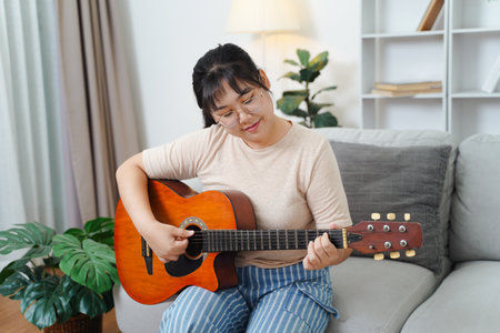 Happy Asian women play acoustic guitar and singing on the sofa in living room at home, enjoying hobbies, relaxation, leisure, and musical concepts.の写真素材