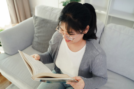 Asian young woman with glasses reading a book on the sofa in the living room happily, seeking knowledge, leisure time, relaxation, hobby concept.の写真素材
