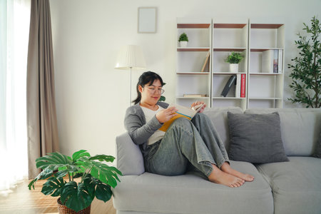 Asian young woman with glasses reading a book on the sofa in the living room happily, seeking knowledge, leisure time, relaxation, hobby concept.の写真素材