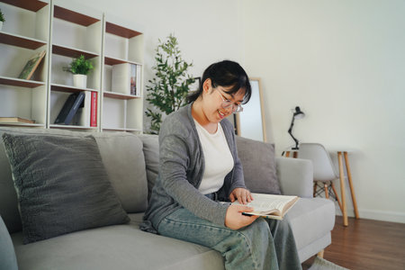 Asian young woman with glasses reading a book on the sofa in the living room happily, seeking knowledge, leisure time, relaxation, hobby concept.の写真素材