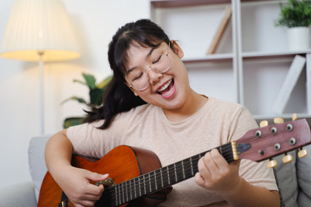 Happy Asian women play acoustic guitar and singing on the sofa in living room at home, enjoying hobbies, relaxation, leisure, and musical concepts.の写真素材