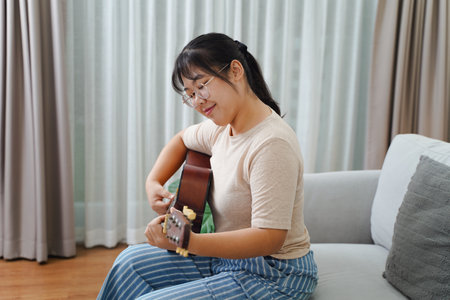 Happy Asian women play acoustic guitar and singing on the sofa in living room at home, enjoying hobbies, relaxation, leisure, and musical concepts.の写真素材