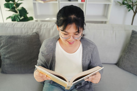 Asian young woman with glasses reading a book on the sofa in the living room happily, seeking knowledge, leisure time, relaxation, hobby concept.の写真素材