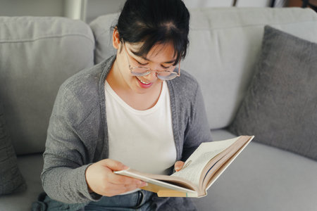 Asian young woman with glasses reading a book on the sofa in the living room happily, seeking knowledge, leisure time, relaxation, hobby concept.の写真素材
