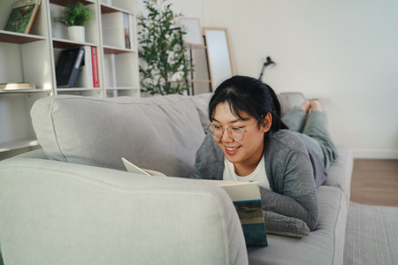 Asian young woman with glasses reading a book on the sofa in the living room happily, seeking knowledge, leisure time, relaxation, hobby concept.の写真素材