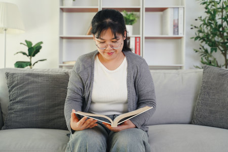 Asian young woman with glasses reading a book on the sofa in the living room happily, seeking knowledge, leisure time, relaxation, hobby concept.の写真素材
