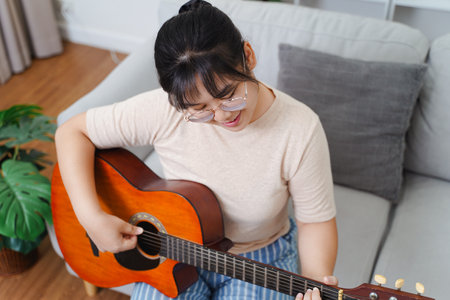 Happy Asian women play acoustic guitar and singing on the sofa in living room at home, enjoying hobbies, relaxation, leisure, and musical concepts.の写真素材