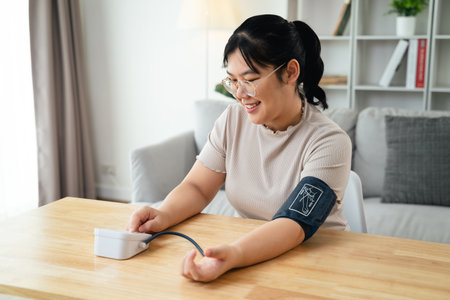 An Asian woman uses a portable blood pressure monitor in her living room. Health awareness concept.