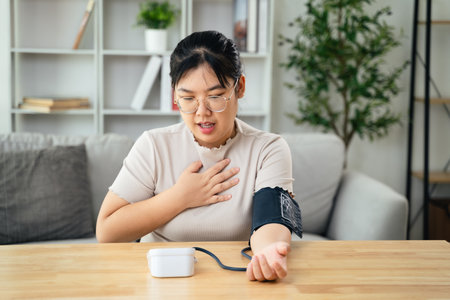 An Asian woman uses a portable blood pressure monitor in her living room, her expression reflecting...
