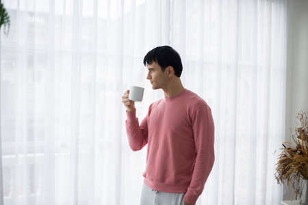 Young man drinking coffee by the window in the room.の写真素材