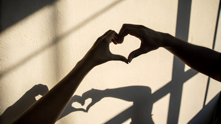 Female hands forming a heart shape with shadow on a white wall.の素材