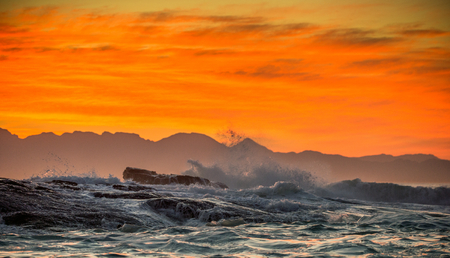 Seascape.  Clouds in a red dawn sky, waves crashing with splashes against stones, silhouettes of...