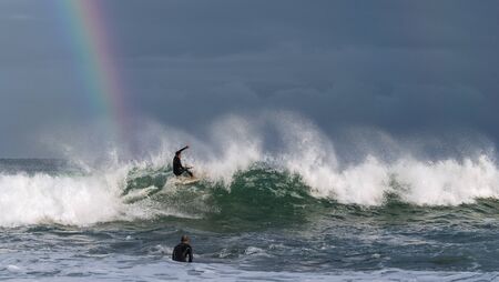 Mossel Bay, South Africa - July 17 2019: Surfing the waves. Surfer riding wave with rainbow on storm sky background.のeditorial素材