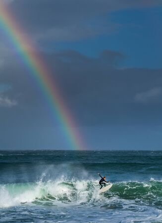 Mossel Bay, South Africa - July 17 2019: Surfing the waves. Surfer riding wave with rainbow on storm...