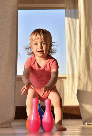 Happy smiling little girl playing bowling with colored pins. In room with cuset light