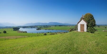 idyllic bavarian landscape with little chapel, lake riegsee and the mountain rangeの写真素材