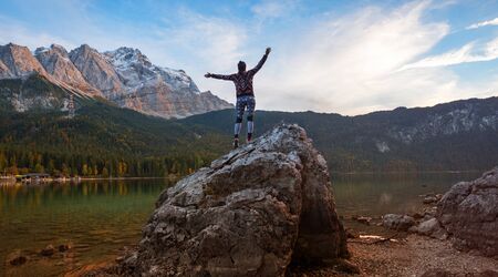 young woman with outstretched arms, doing gymnastics on a big rock at eibsee lake shore. evening...