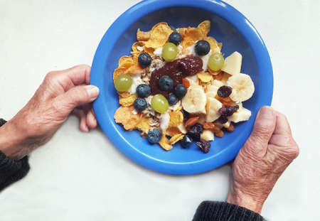 breakfast for seniors. old hands holding blue plate with cereal and fresh fruitsの写真素材