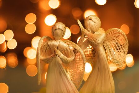 Two straw angels praying on a blurred Christmas background. Beautiful bokeh created by Christmas lights. Traditional hand-made doll as a Christmas tree decoration.の写真素材