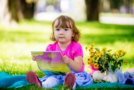 Portrait of little smiling girl child with book sitting on the grass in sunny summer day.の写真素材