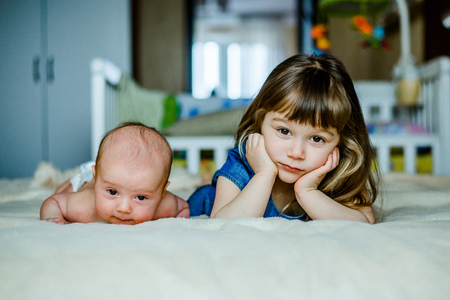 Cute little girl is playing with his younger brother at home lying on the bed with cupsの写真素材