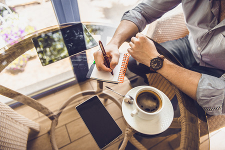Businessman in a cafe drinking coffee, writing in a notebook, the tablet on the tableの写真素材