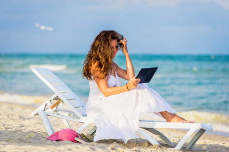 Young woman in hat with tablet pc at the beach. girl sitting on a deck chair with the tabletの写真素材