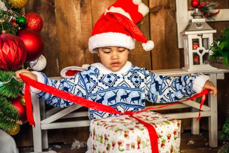 Little boy opening a Christmas present. African-American boy in the hat of Santa Claus opens...