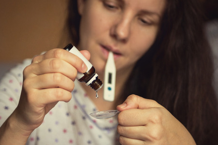 Woman with a thermometer in his mouth dripping curative medicine in a spoonの写真素材