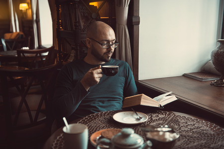a young man with glasses, beard, sitting in cafe and reading a bookの写真素材