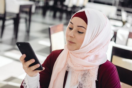 Muslim girl dials a number on a cell phone, sitting in a cafe with a cup of coffeeの写真素材