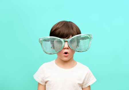 Cheerful little boy in big glasses express a surprised face isolated on blue backgroundの写真素材