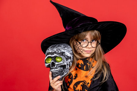 Handsome child dressed in black dress for Halloween carnival with a human skull in his hands. The girl is isolated on a red background in a witch costume.の写真素材