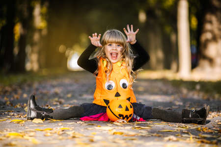 A girl in a stylish pumpkin costume and a hat yells and gestures while sitting on a twine while celebrating Halloween in an autumn parkの写真素材