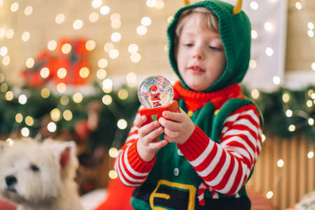 Happy new year concept. Happy blond child sits on the bed, opens a box and takes out a gift glass snow globe, the child smiles happily and is pleased with the gift.の写真素材