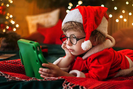 Cute boy in Santa Claus costume watching Christmas cartoons on tablet, preparing for celebration, wonderful atmosphere and decorations, miracleの写真素材