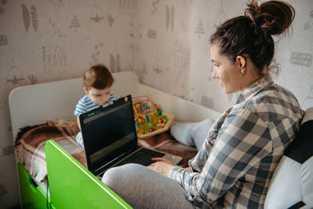 cute baby 6 months old European appearance sits on bed with mom working on maternity leaveの写真素材