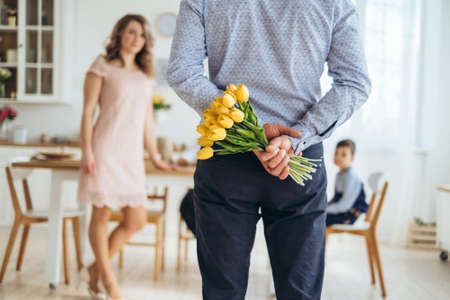 A young man holds yellow tulips behind his back. Surprise gift for the girl on March 8, Valentines Day. Flowers in the hands of a man in a blue shirt.の写真素材
