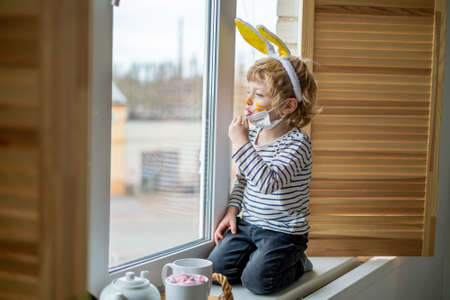 Easter 2021 concept: A child in home quarantine plays by the window with the ears of a rabbit on his...
