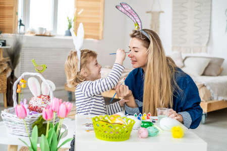 Beautiful happy son and mother in headbands with bunny ears decorate eggs with a brush and bright paints, prepare to celebrate Easter, have fun at homeの写真素材