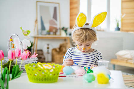 Cute little kid boy with bunny ears headband and face make up coloring eggs for Easter holiday with paint and brush, decorating eggs, developing creativity and imagination, happily spending time homeの写真素材