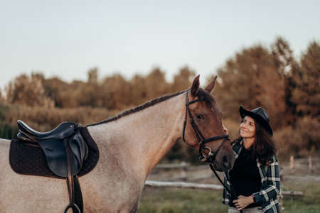Beautiful smiling young woman in hat and shirt playing with brown horse in a field at sunset. Horseback riding.の写真素材