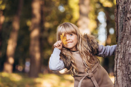 Beautiful blonde girl covers one eye with yellow maple leaf outdoors in autumn park.の写真素材