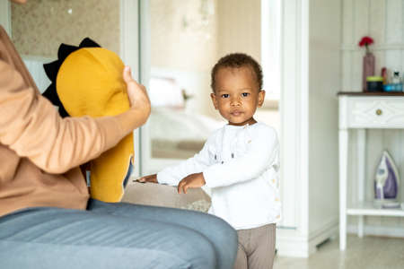 Loving mommy showing cute dino hat to her sweet afro-american son, friendly family preparing going for a walk, choosing clothes together, upbringing psychologyの写真素材