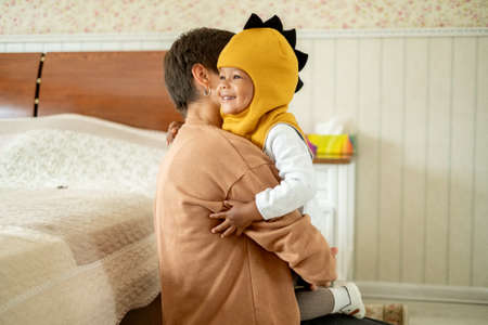 Loving mommy showing cute dino hat to her sweet afro-american son, friendly family preparing going for a walk, choosing clothes together, upbringing psychologyの写真素材